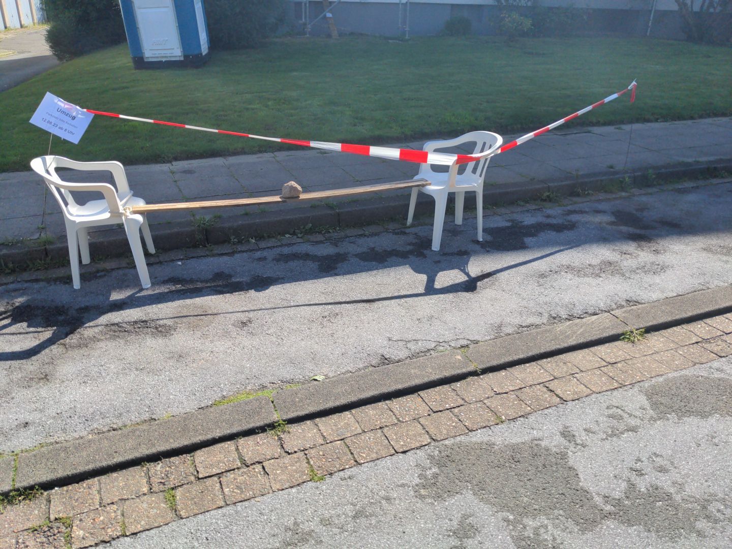auf einer Straße stehen zwei Plastikstühle. dazwischen ist eine Holzplanke aufgehängt. auf der Planke liegt ein Stein. darum ist absperrband und ein ausgedruckter uns laminierter Zettel mit "Umzug, 12.08.2023 ab 8 Uhr"